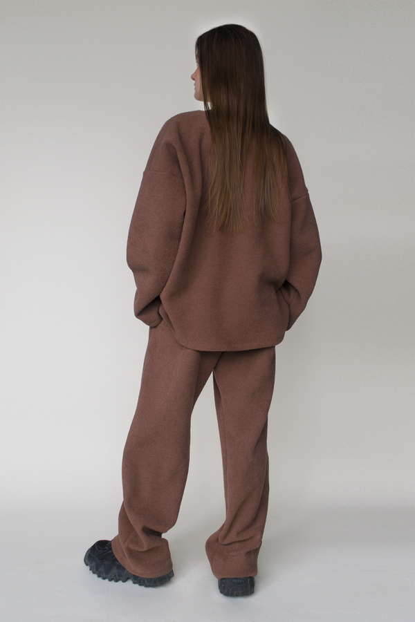 Woman standing in front of a grey backdrop, wearing a brown oversized large fleece sweater, and brown fleece pants. The outfit is a unisex style, paired with black sneakers. She has long brown hair and fair skin.