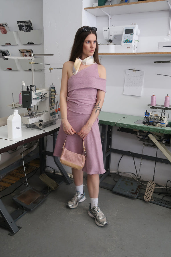 young woman standing in fashion studio wearing a mauve one-shoulder tank top, with a band that crosses over the chest and arm. Design by Alex Watson Studio. Paired with a silk scarf, little purse and sunglasses.
