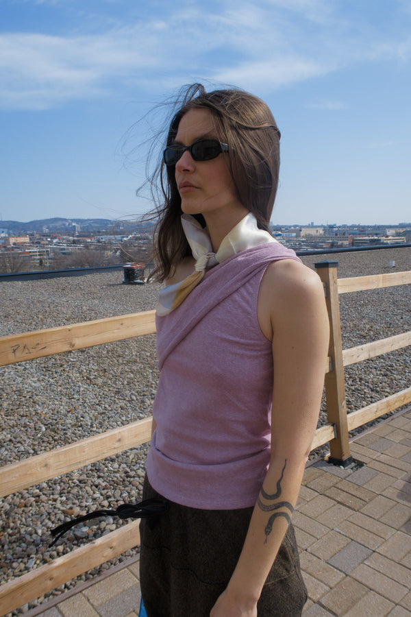 young woman standing in fashion studio wearing a mauve one-shoulder tank top, with a band that crosses over the chest and arm. Design by Alex Watson Studio. Paired with a silk scarf, little purse and sunglasses.