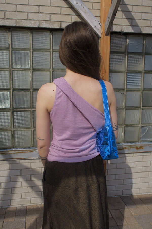 young woman standing outside wearing a mauve one-shoulder tank top, with a band that crosses over the chest and arm. Design by Alex Watson Studio. Paired with a silk scarf, little purse and sunglasses.
