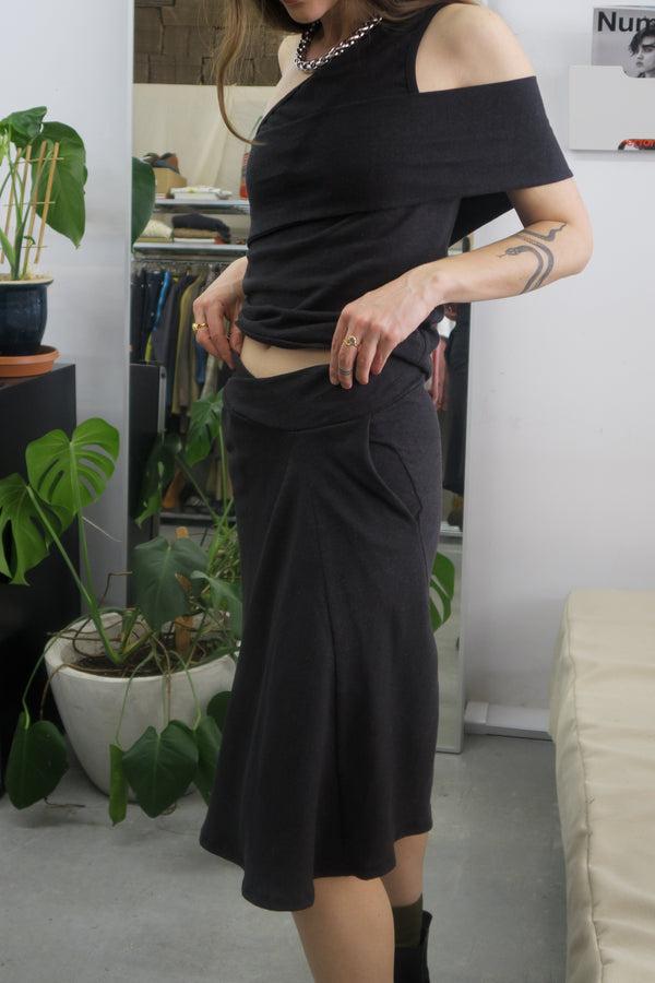 Woman standing in fashion studio wearing a black skirt and matching top. Paired with a silver necklace, and black boots. The skirt has a crossover waistband and fits to the knees.