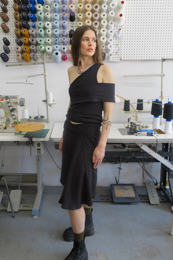 Woman standing in fashion studio wearing a black skirt and matching top. Paired with a silver necklace, and black boots. The skirt has a crossover waistband and fits to the knees.