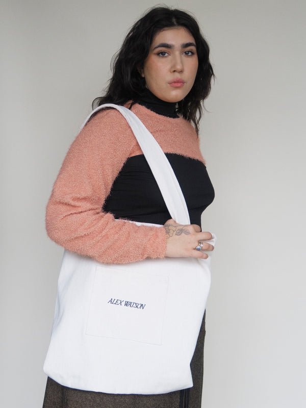 woman standing carrying a heavyweight canvas totebag with Alex Watson embroidered onto the front pocket. She's wearing a cropped orange shrug sweater and black tank top. She has long black hair