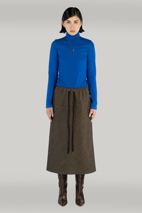 Young woman with short black hair stands confidently wearing a cobalt blue turtleneck and long brown wool skirt with a bias cut. She completes her look with brown boots and gold necklace