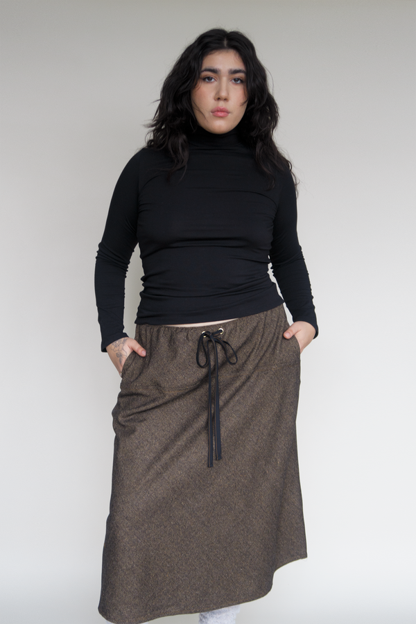 Woman standing with long black wavy hair, plus size. wearing black fitted turtleneck and long brown wool skirt and black drawstring tied in a bow.